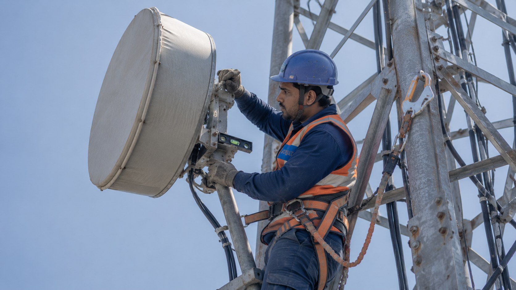 Rigger aligning a microwave dish antenna at height on a telecom tower, alignment tool in hand, harness clipped to the structure.